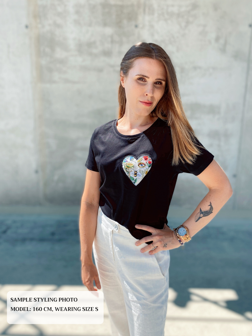 Woman wearing a black t-shirt with a colorful heart design embroidery, standing against a concrete wall.