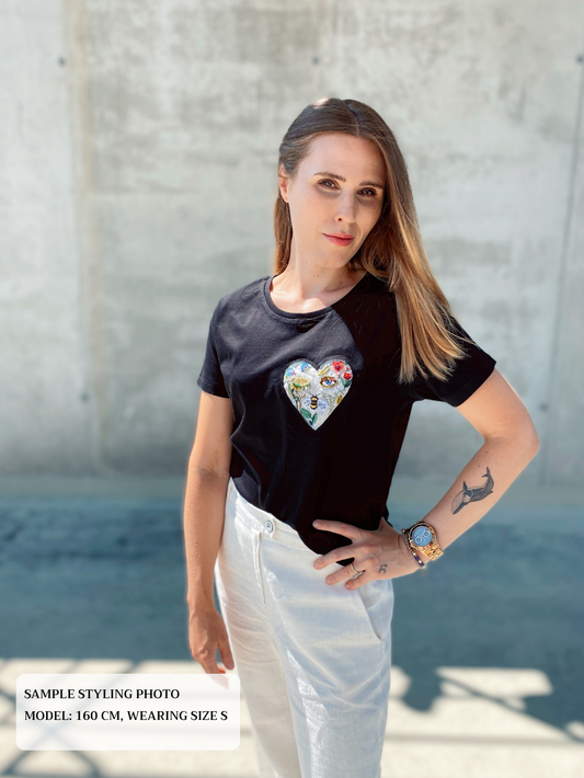Woman wearing a black t-shirt with a colorful heart design embroidery, standing against a concrete wall.