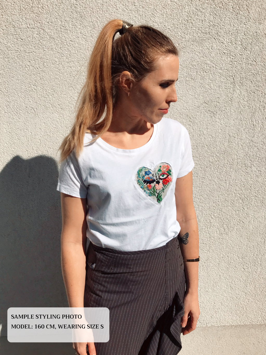 Woman wearing a white t-shirt with a colorful heart embroidery design against a light wall.