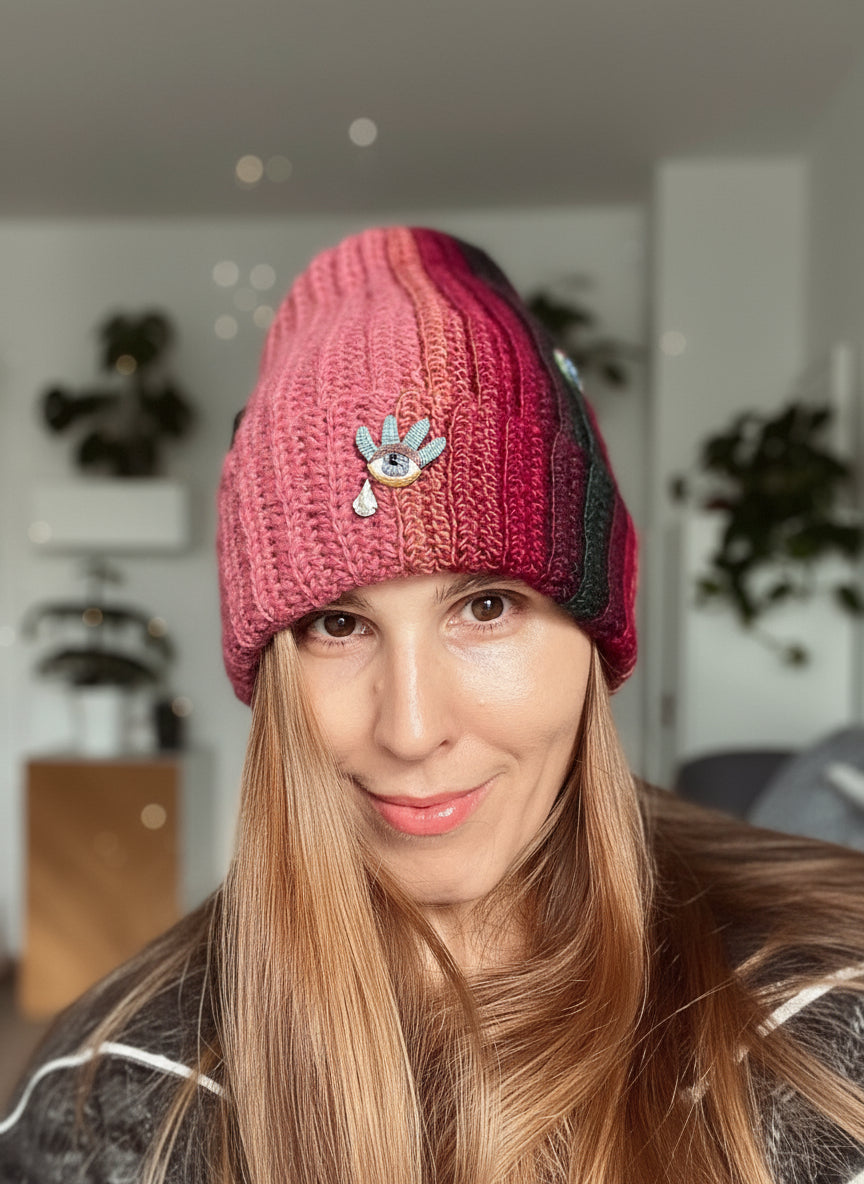 Person wearing a pink knitted beanie with a decorative flower emblem indoors.