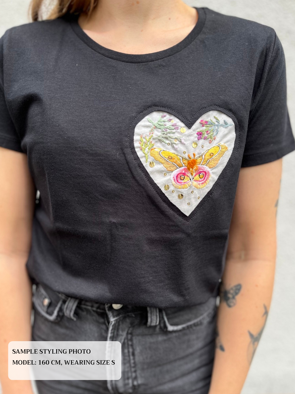 Close up of a woman wearing a black t-shirt with a heart-shaped embroidered design featuring a moth and flowers.