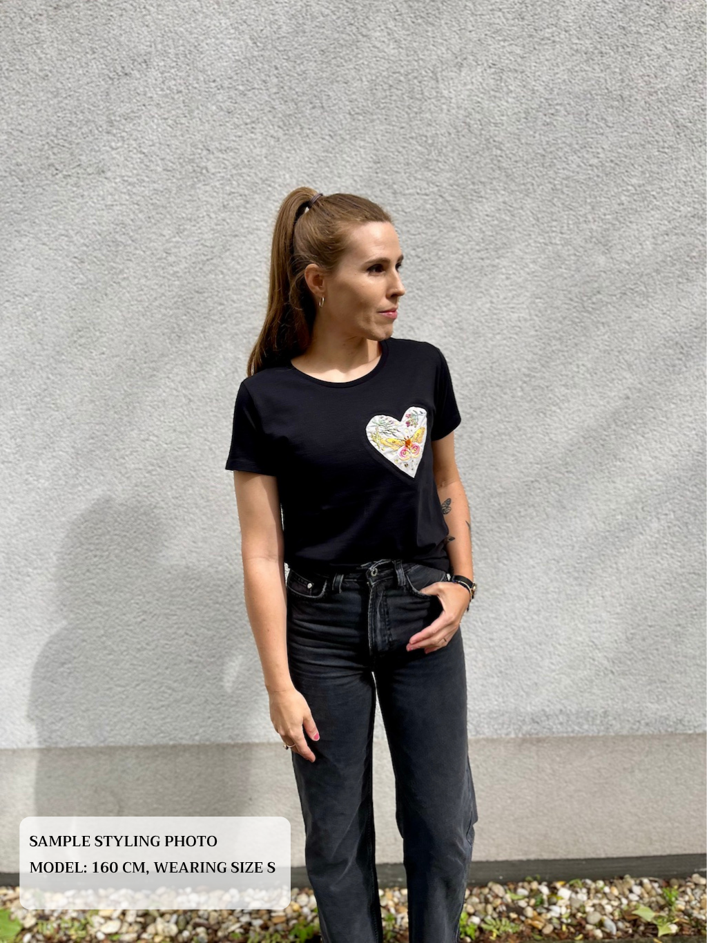 Woman wearing a black t-shirt with embroidered heart design detail and dark jeans against a gray wall.