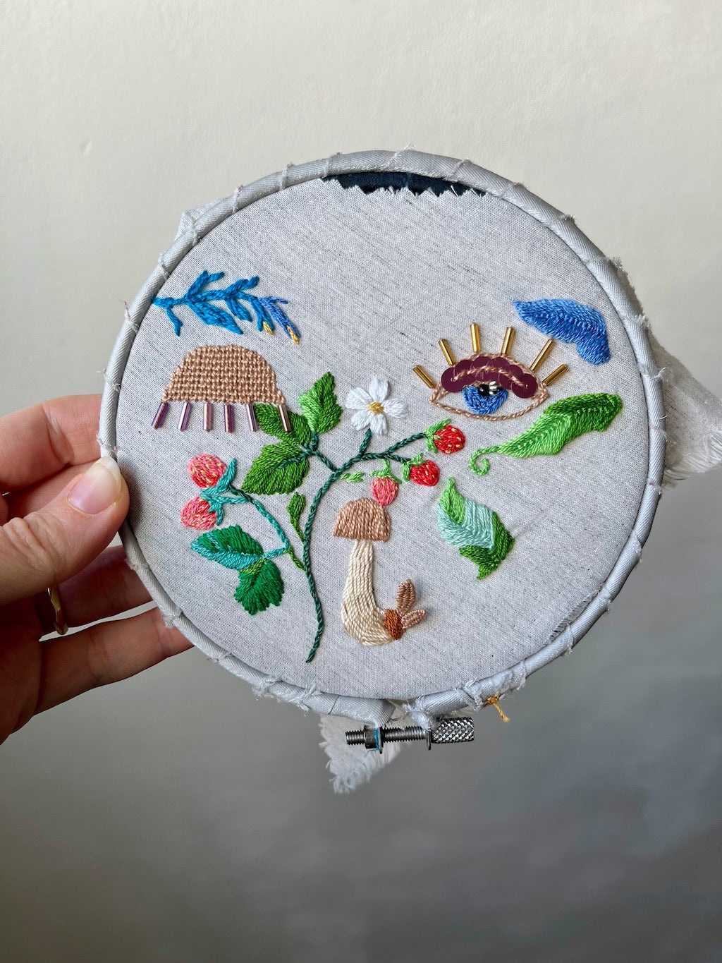 Embroidery hoop with strawberries and mushroom design held by a hand against a neutral background.