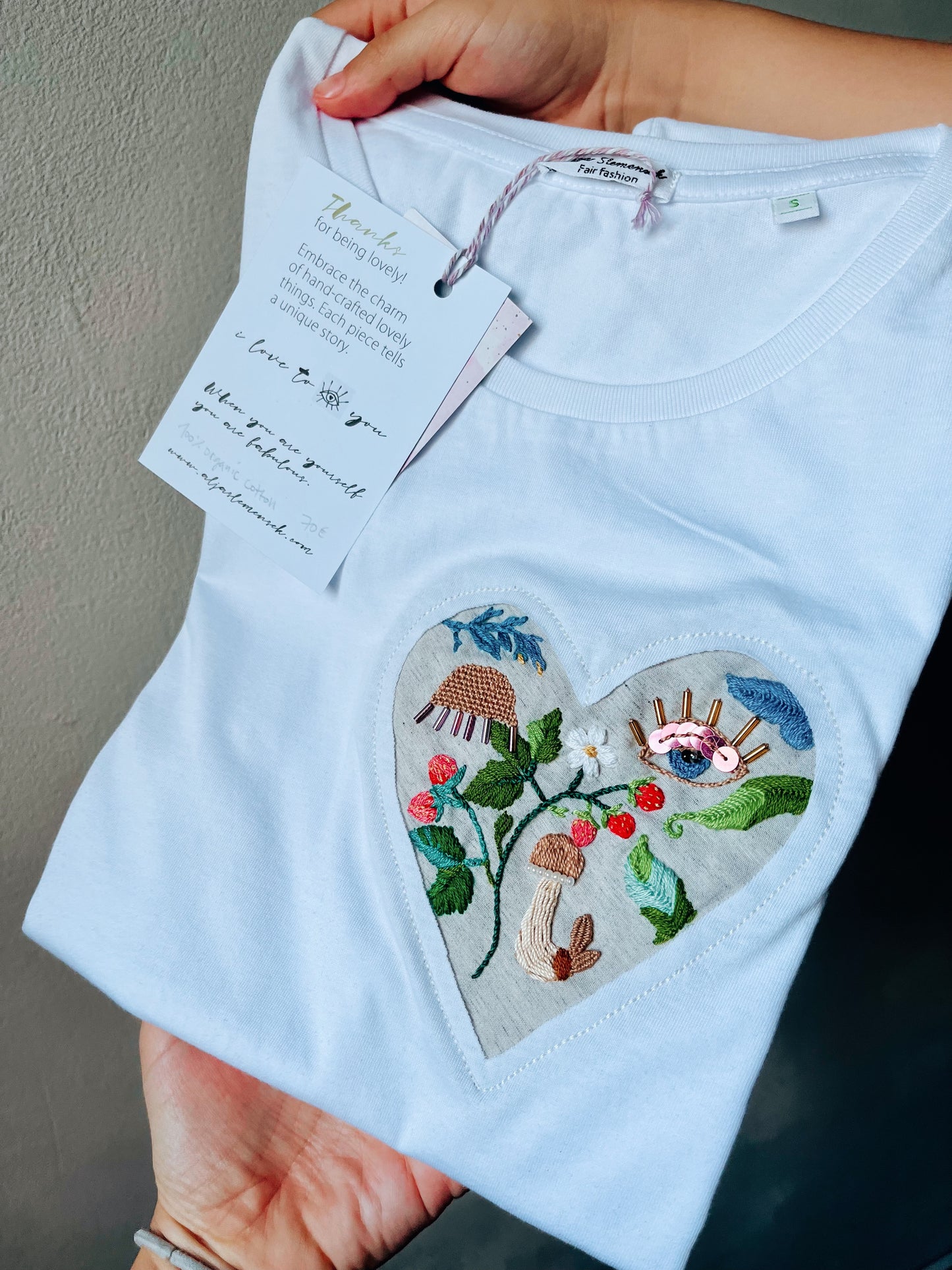 White t-shirt with a heart-shaped embroidered design held by a woman.