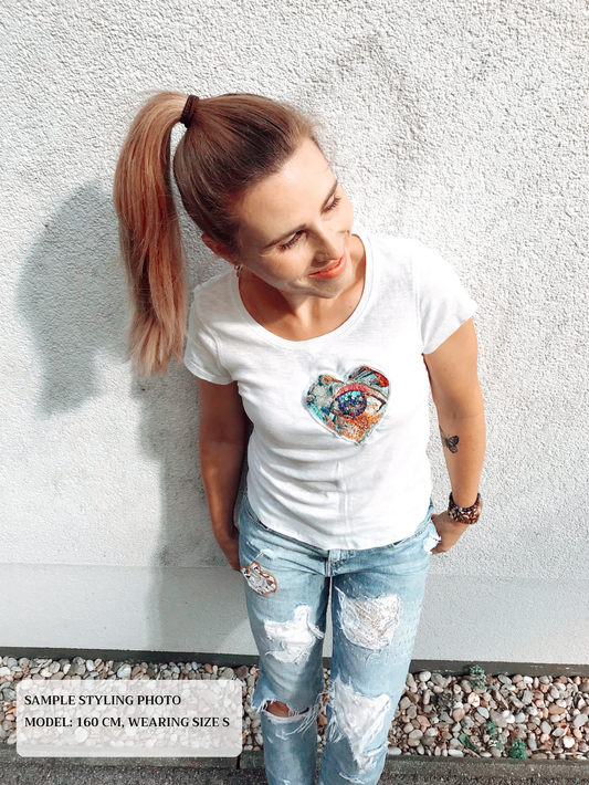 Woman wearing a white t-shirt with a colorful embroidery design and ripped jeans against a textured wall.