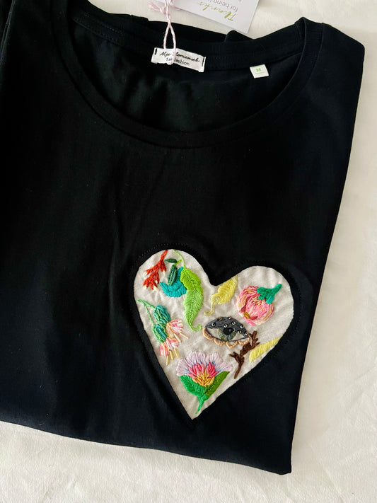 Black t-shirt with a colorful heart-shaped embroidery featuring wildflowers and green leaves on a white surface.